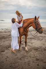 Horse riding on the beach. Cute little girl on a brown horse. Her mom standing near by. Love to animals. Mom and daughter spending time together. Outdoor activities. Mother's day.