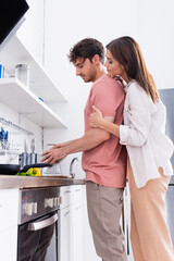 Side view of woman hugging boyfriend holding grater near vegetables in kitchen