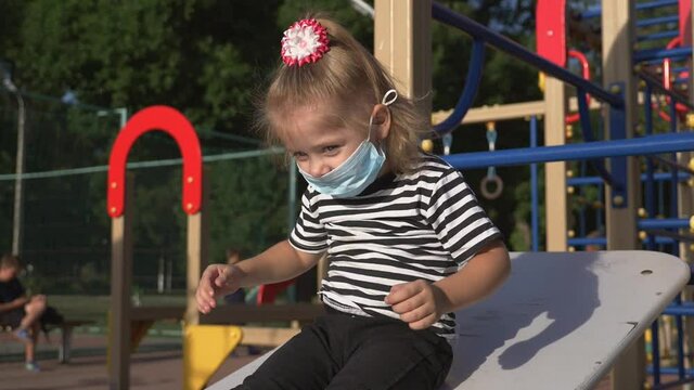 little girl wears a medical safety mask on his face. kid is playing outside in children's park. concept of kid safety from contracting an infection. girl is protected in a safety mask in park.