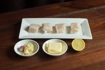 Food plating and presentation of raw food on an old wooden table. Hake, butter, garlic, and half a lemon.