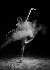 A beautiful barefoot slender ballet dancer girl wearing a bodysuit, posing dancing among the clouds of flying flour on a black background. Artistic, commercial, monochrome design