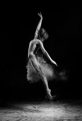 A beautiful barefoot slender ballet dancer girl wearing a bodysuit, dances and jumps among the clouds of flying flour on a black background. Artistic, commercial, monochrome design