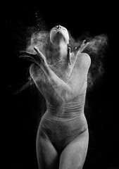 A beautiful slender ballet dancer girl wearing a bodysuit, sensually poses among the flying flour which covers her body, on a black background. Artistic, commercial, monochrome design