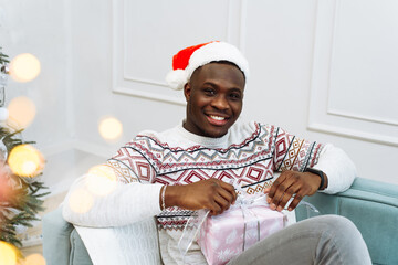 Portrait of a handsome young black man opening a present in a Christmas hat on a sofa with a Christmas tree in the background