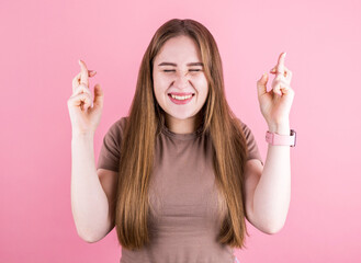 Closeup of happy lovely young woman wears t shirt keeps fingers crossed and looks up, isolated over pink background