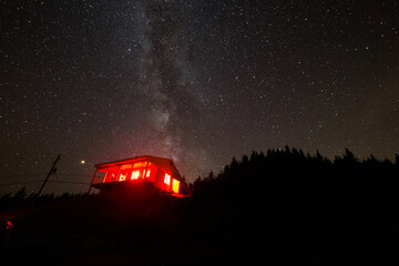 petite maison avec une lumi&egrave;re rouge au sommet d'une montagne avec la voie lact&eacute;e en arri&egrave;re plan
