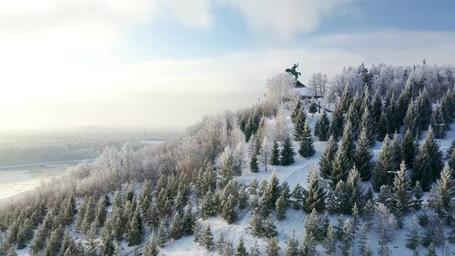 Drone shot of Monument to Salavat Yulaev on a mountain among rows of firs in sunny cold winter day with beautiful cityscape and river on background. High quality 4k footage.