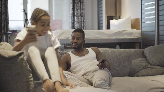 Joyful teenage girl joining father watching TV and showing video recorded for social media. portrait of friendly African American family enjoying weekends at home.