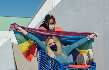 Two women with mask playing with a shopping cart and a lgtbflag