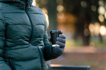 The guy is holding a paper cup with coffee or tea in winter gloves. Have a cup of coffee. Disposable paper cup. Hot drink. Empty space. Man standing on the street holding a cup of coffee. Cup.