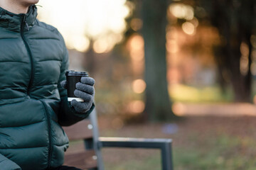 The guy is holding a paper cup with coffee or tea in winter gloves. Have a cup of coffee. Disposable paper cup. Hot drink. Empty space. Man standing on the street holding a cup of coffee. Cup.