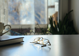 In front of the window on the table are glasses, a diary and a cup of coffee. Solar illumination. Concept-work at home