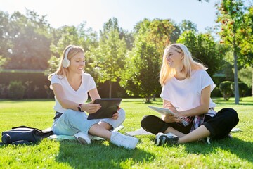 Girls teenage students sitting on green lawn in park with backpack, digital tablet