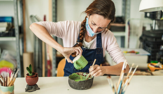 Mature woman works with ceramics and paints finished cup while wearing protective face mask for coronavirus - Powered by Adobe
