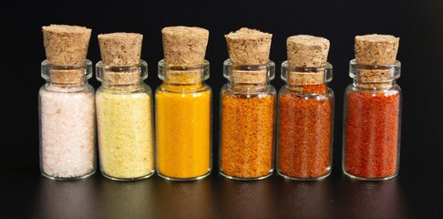 A set of spices in small jars with cork lids. Pink salt, ginger, turmeric, masala, paprika, smoked paprika. Spices on a black surface