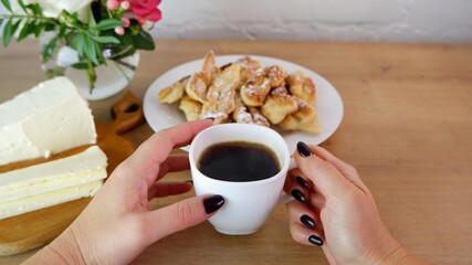 Cup of coffee in female hands, close-up. Homemade baked goods during quarantine. Homemade feta cheese on a wooden tabletop. Concept on the background of a white brick wall and a bouquet of roses.