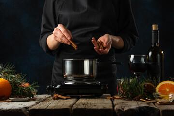 Professional chef pours cinnamon stick into pan for preparing mulled wine on rustic wooden table with festive composition background. Backstage of cooking hot drink with fragrant spices.