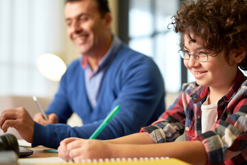 Fototapeta premium Portrait of smiling teenaged latin boy in glasses making notes while sitting at the desk together with his father and doing homework at home