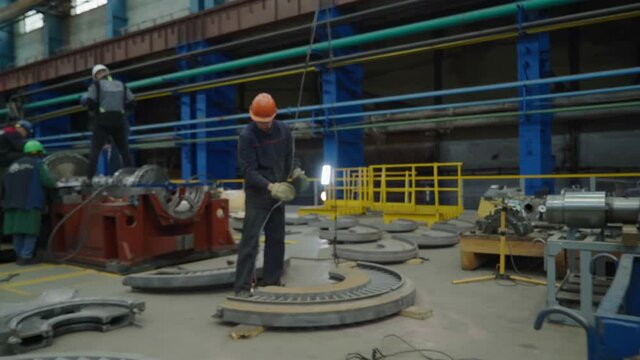 Gas and steam turbine production plant. A worker is preparing a big metal detail for further assembly. Unfastening the crane straps and placing it on the ground. Manual labor. Heavy industrial plant