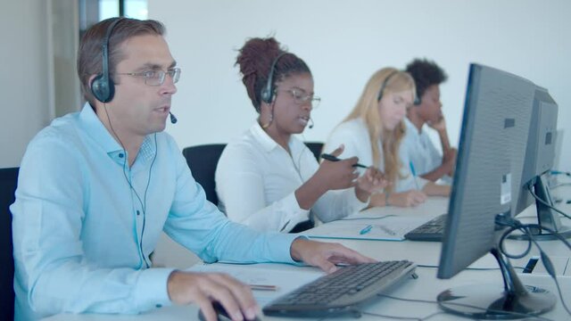 Team of call center consultants in headset sitting in row, using computer while talking to customer. Customers assistance or telemarketing concept.