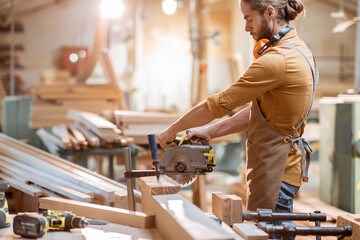 Carpenter sawing wooden bars with cordless electric saw at the joiner's workshop