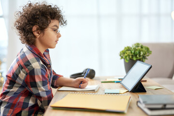 Get an education. Enthusiastic latin school boy wearing glasses making notes, having online lesson using digital tablet while studying at home