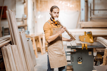 Handsome carpenter in uniform working with wood, checking the quality of the wooden baluster at the...