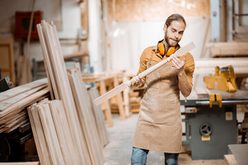Handsome carpenter in uniform working with wood, checking the quality of the wooden baluster at the...