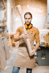 Handsome carpenter in uniform working with wood, checking the quality of the wooden baluster at the joinery