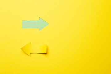 Two opposite left/right arrows, one cutted from the yellow paper curved up of two sides on the yellow paper background other made as an arrow shaped hole in the background with green paper underlay