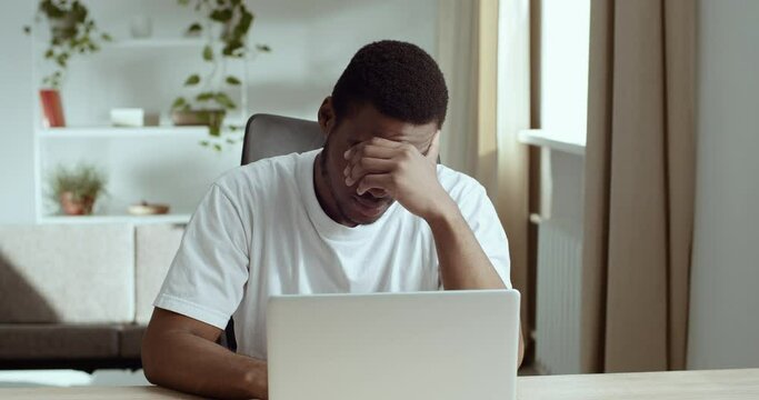 Portrait of afro american student black male tired guy wears white looks at laptop screen learns bad new feels exhaustion stress worry suffers from headache holding his head with hand, problem concept