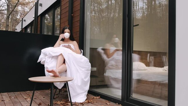 Young brunnete woman relaxing in forest house. The naked lady is wrapped in blanket and having a cup of coffee enjoying time alone.