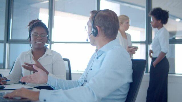 Two cheerful sales agents in headsets sitting together and talking to each other, discussing calls and smiling. Teamwork or telemarketing concept.