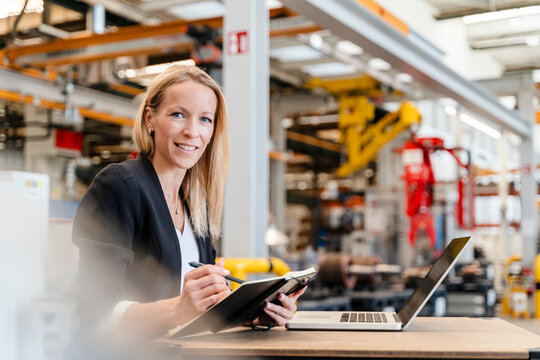 Smiling businesswoman holding diary while sitting with laptop in industry