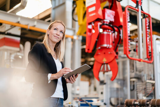 Smiling blond businesswoman looking away while holding digital tablet standing in factory
