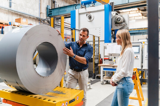 Businessman explaining female colleague about steel roll while standing in factory