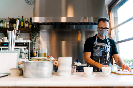 Male barista wearing protective face mask while working in kitchen of coffee shop during COVID-19 - Powered by Adobe