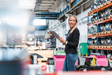 Happy female entrepreneur holding digital tablet looking away while standing in factory