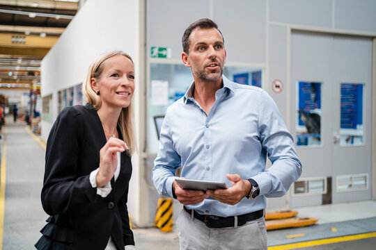 Smiling female entrepreneur and businessman looking away while holding digital tablet standing at industry