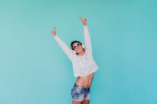 Happy fashionable woman listening music while standing with arms raised against turquoise background