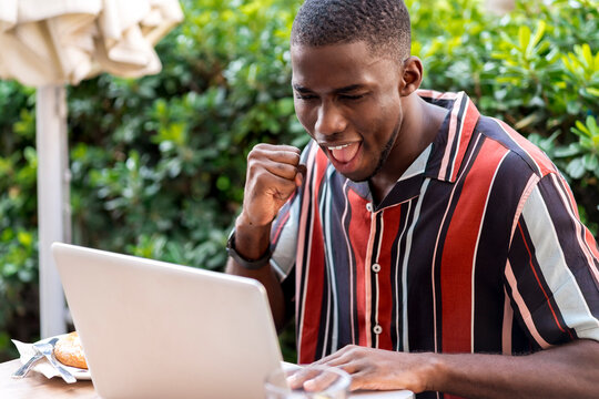Happy young man clenching fist while using laptop on terrace of cafe