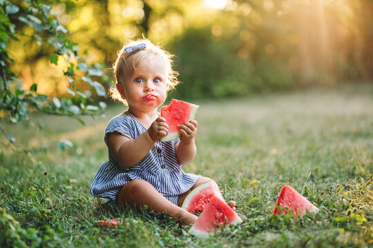 Cute Caucasian Baby Girl Eating Ripe Red Watermelon In Park. Funny Child Kid Sitting On Ground With Fresh Fruit Outdoors. Supplementary Healthy Finger Food For Toddler Kids. Summer Fun.