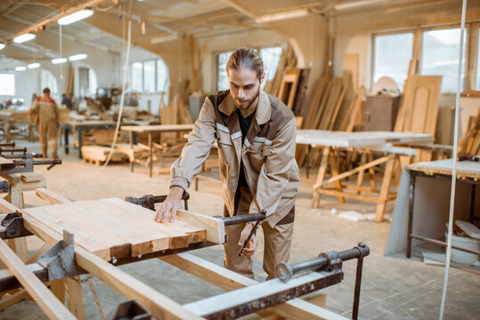 Handsome carpenter in uniform gluing wooden bars with hand pressures at the carpentry manufacturing