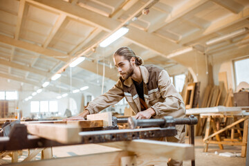 Handsome carpenter in uniform gluing wooden bars with hand pressures at the carpentry manufacturing - obrazy, fototapety, plakaty