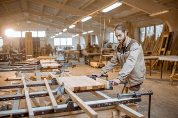 Handsome carpenter in uniform gluing wooden bars with hand pressures at the carpentry manufacturing - obrazy, fototapety, plakaty