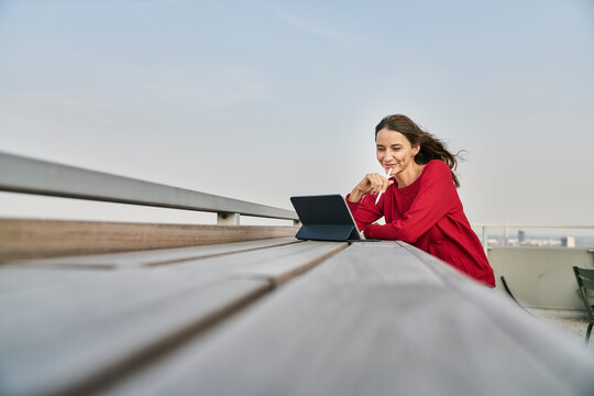 Smiling businesswoman holding digital tablet while leaning on retaining wall at rooftop