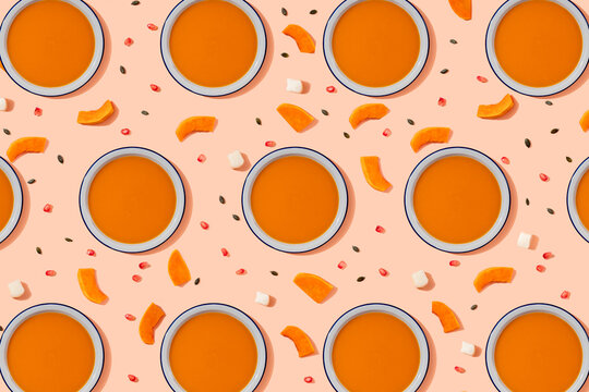 Pattern of bowls of pumpkin soup surrounded by pumpkin seeds, pomegranate seeds and pieces of cheese and pumpkin