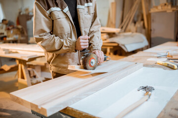 Handsome carpenter in uniform brushing wood with hand machine at the carpentry manufacturing