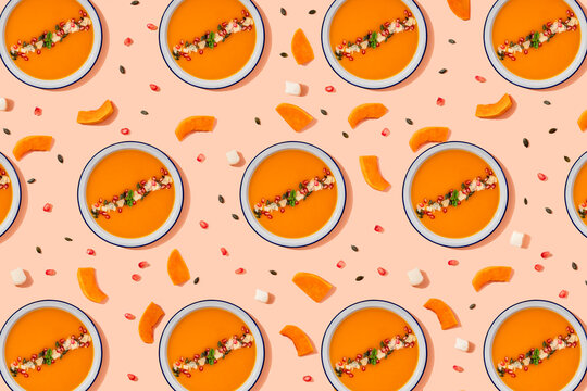 Pattern of bowls of pumpkin soup surrounded by pumpkin seeds, pomegranate seeds and pieces of cheese and pumpkin