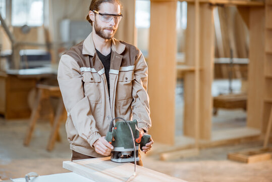 Handsome carpenter in uniform chamfers wooden bar with a hand machine at the carpentry manufacturing